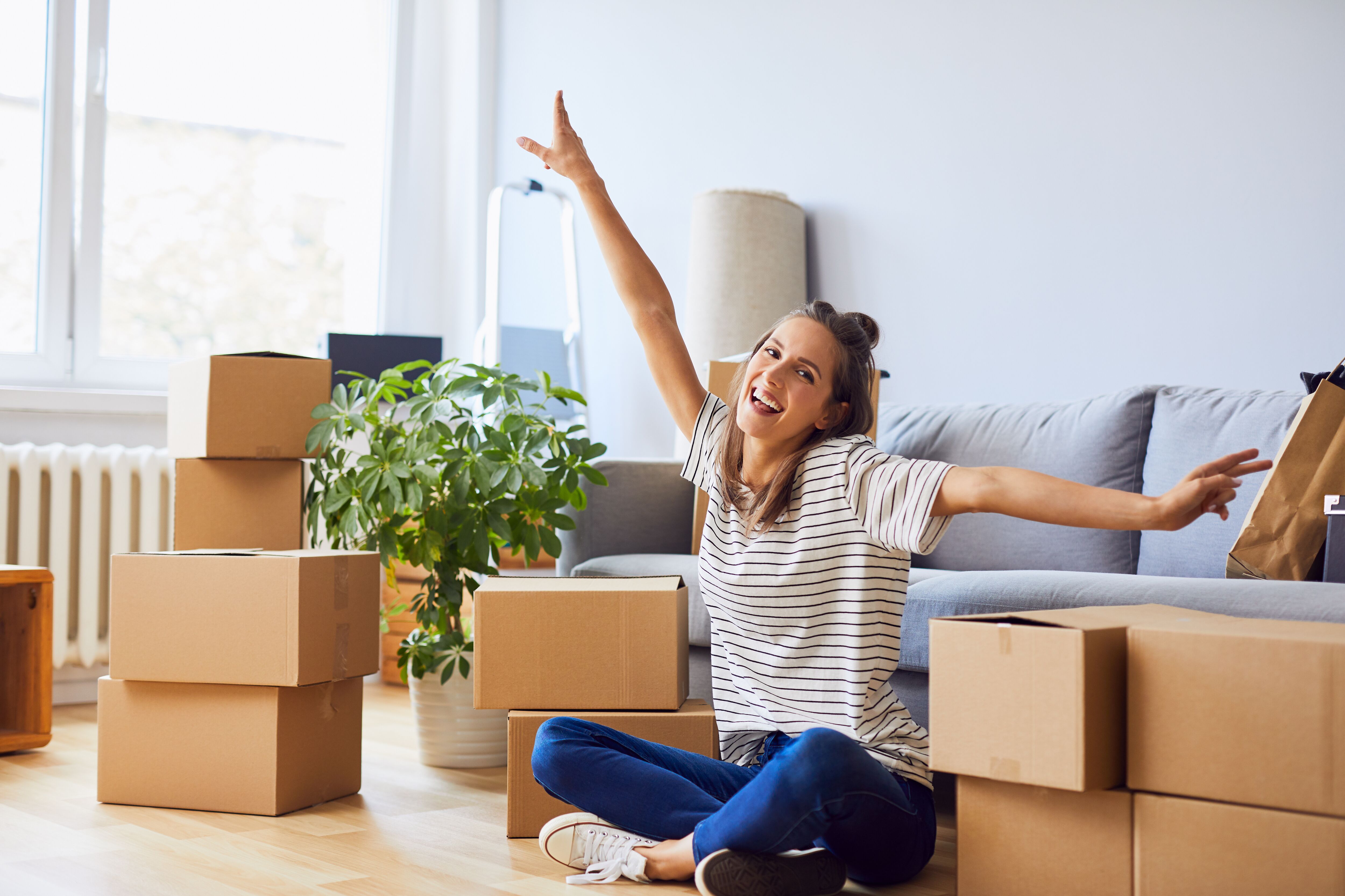 Young woman sitting in new apartment and raising arms in joy after moving in.