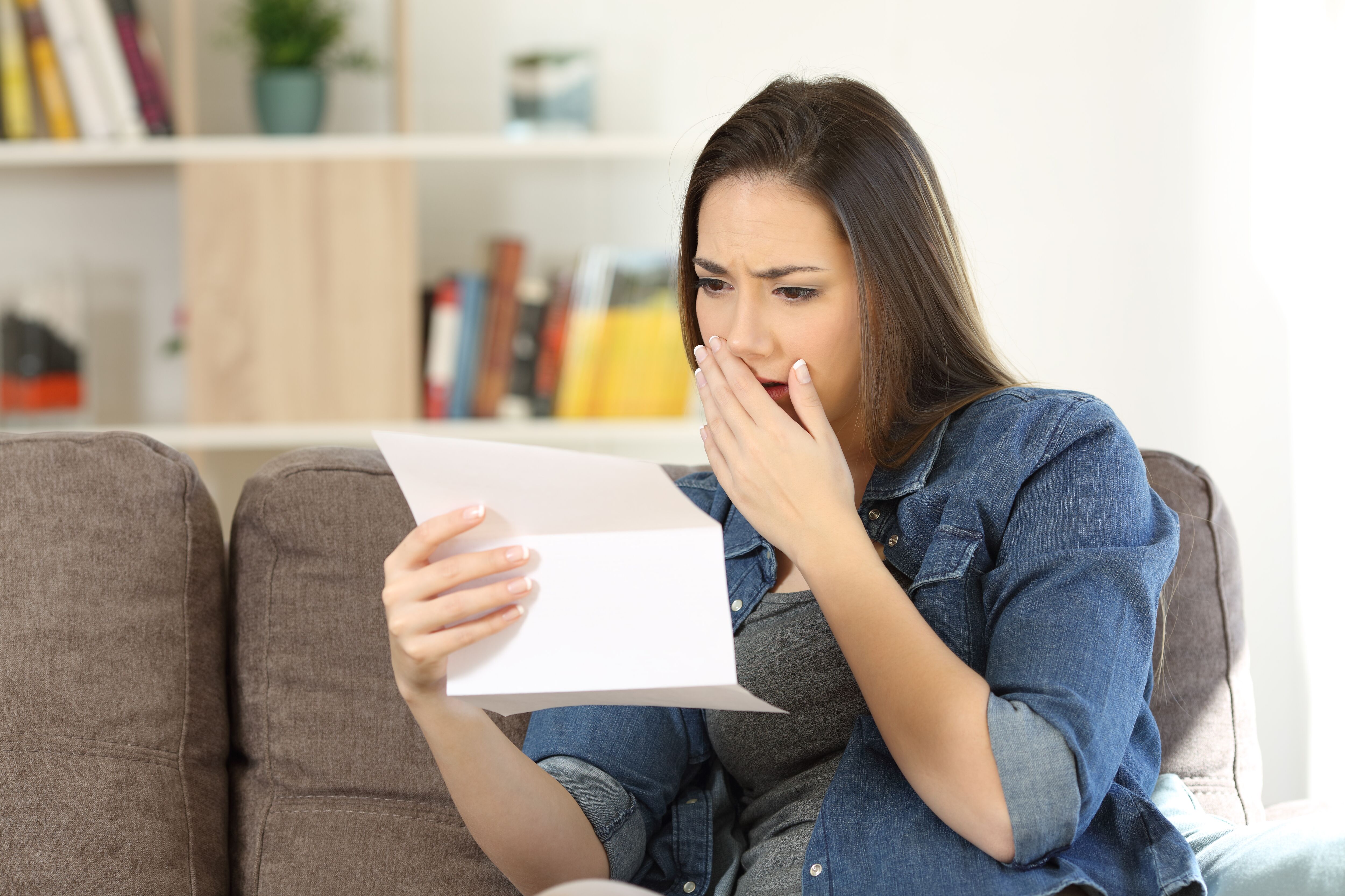 Sad woman reading bad news in a paper letter sitting on a couch in the living room at home