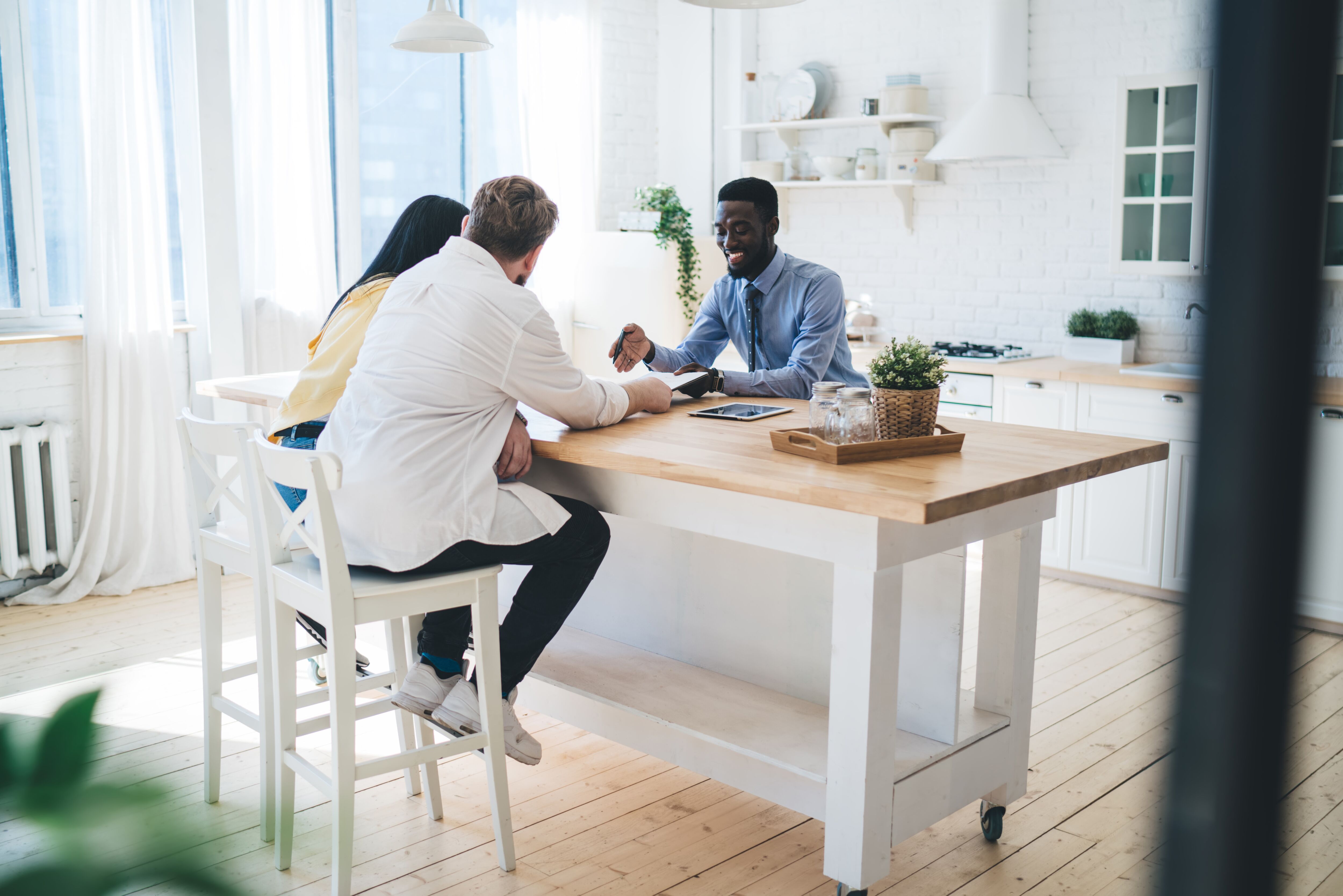 Cheerful and friendly black realtor sitting in bright kitchen and explaining details of contract to couple of clients while smiling